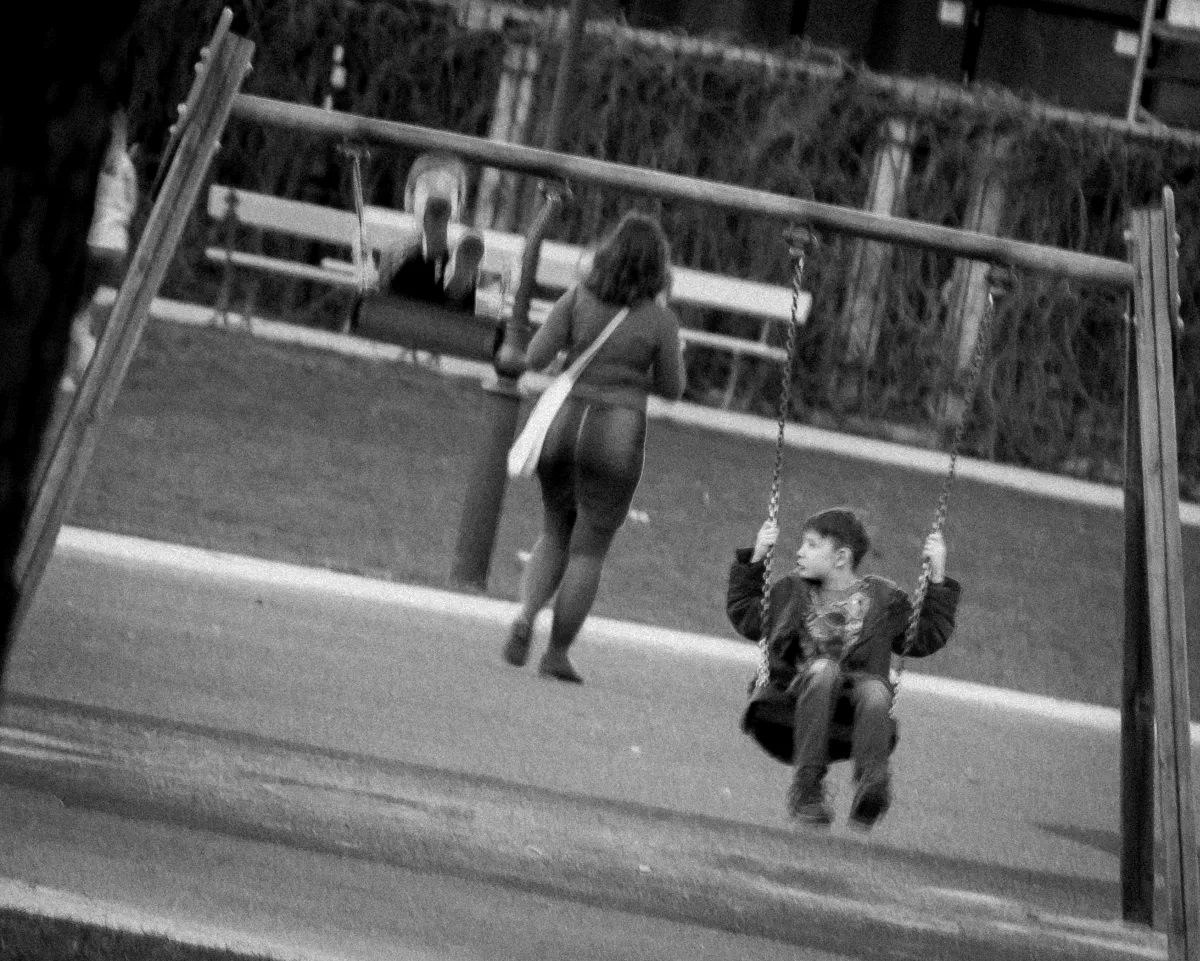Child on a playground swing looking sideways while a woman walks away